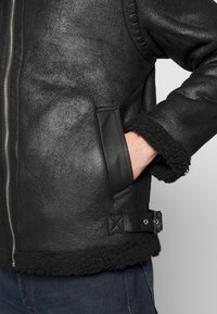 Giacca in shearling nera con finitura in pelle testurizzata, tasche laterali, polsini foderati in pile e dettaglio con fibbia in vita.