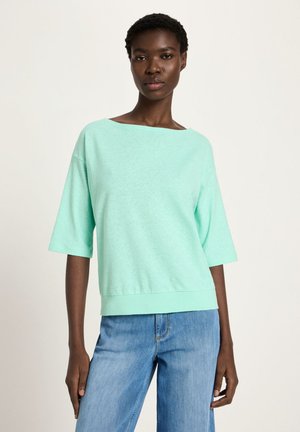 Frau, die ein locker sitzendes, mintgrünes Kurzarmshirt und hellblaue Jeans trägt, steht vor einem schlichten hellen Hintergrund.