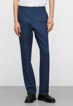 Man draagt slanke marineblauwe broek met zwarte leren loafers, staande tegen een effen lichte achtergrond.