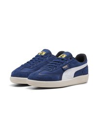 Blå ruskinds Puma Palermo sneakers med hvid sidebånd, beige sål og Ferrari-logo på tungen, vist mod hvid baggrund.