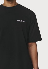 Černé bavlněné tričko s krátkým rukávem, které na levé části hrudi má bílé vyšívané logo "PEGADOR".