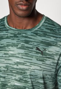 Roheline mustriga sportlik T-särk ümmarguse kaelusega, millel on horisontaalsed jooned, hingav tekstuur ning must Puma logo rinnal.