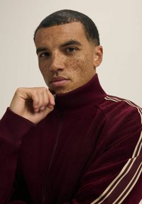 Bordo kurtka na zamek z wysokim kołnierzem; posiada fakturowany materiał i kontrastowe kremowe paski na rękawach. Zbliżenie modelki trzymającej kołnierz.