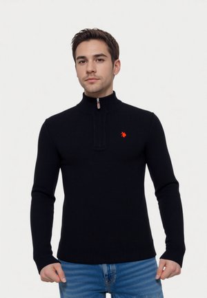 Donkerblauwe gebreide trui met een halve ritssluiting en geribde manchetten. Heeft een klein rood logo op de linkerbuste. Draagt met een blauwe spijkerbroek.
