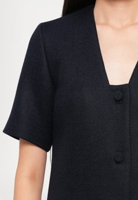 Sötétkék textúrájú ruha V-nyakkivágással, rövid ujjal és egy kör alakú gomb részlettel az elején.