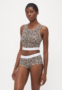 Žena stojí v leopardom vzore bezrukávovom crop topi a zodpovedajúcich šortkách na vysoký pás s bielymi elastickými pásmi.