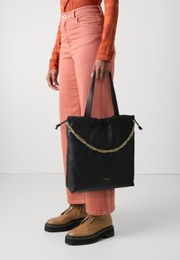 Sort læder tote taske med snøre lukning, prydet med en guld kæde detalje og flade håndtag, holdt af en person iført orange og pink påklædning.