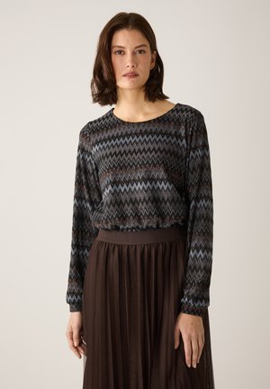 Langärmliges Oberteil mit Zickzackmuster in Dunkelblau, Schwarz und Kupfer; kombiniert mit einem plissierten braunen Rock mit tailliertem Bund.