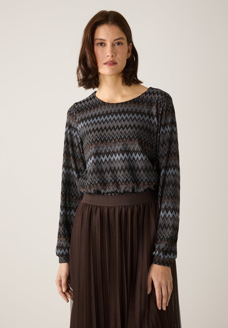 Langärmliges Oberteil mit Zickzackmuster in Dunkelblau, Schwarz und Kupfer; kombiniert mit einem plissierten braunen Rock mit tailliertem Bund.