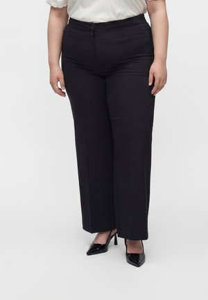 Personne grande taille portant un pantalon noir taille haute, des talons noirs à bout pointu, et un chemisier blanc à manches courtes.