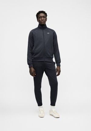 Uomo in piedi rivolto in avanti indossa una giacca sportiva scura con zip, pantaloni abbinati, calzini bianchi e sneakers beige su uno sfondo chiaro e uniforme.