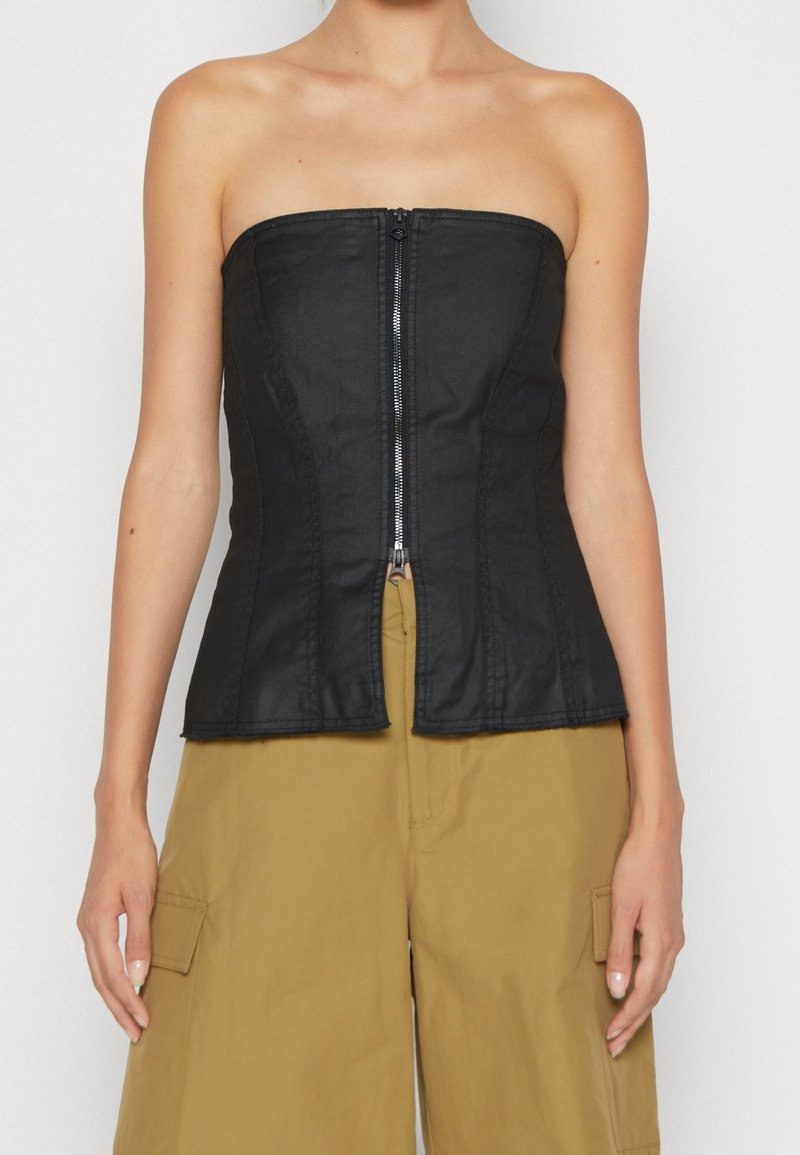 Femme portant un corset noir sans bretelles à fermeture éclair, associé à un pantalon cargo ample couleur moutarde, sur fond uni.