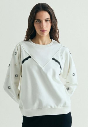 Joven mujer con cabello largo lleva una sudadera blanca con patrones decorativos negros de sol y círculos en las mangas y el pecho.