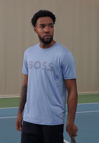 Camiseta atlética de color azul claro con el logo "BOSS" en un tono más oscuro, cuello redondo, mangas cortas, complementada con pantalones cortos negros, de pie en una cancha de tenis.
