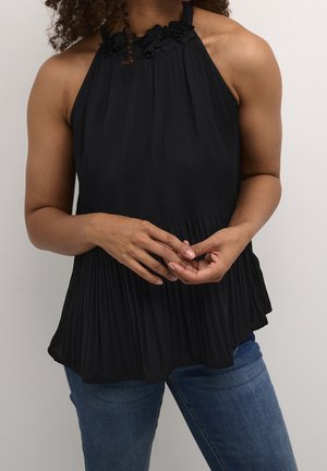 Vrouw draagt een mouwloos zwart geplooid haltertop met bloemrijke halslijn en een blauwe spijkerbroek, handen losjes in elkaar gevouwen voor haar.