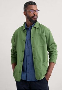 Green button-up shirt jacket in textured fabric, featuring a chest pocket and rolled sleeves, layered over a blue striped shirt.