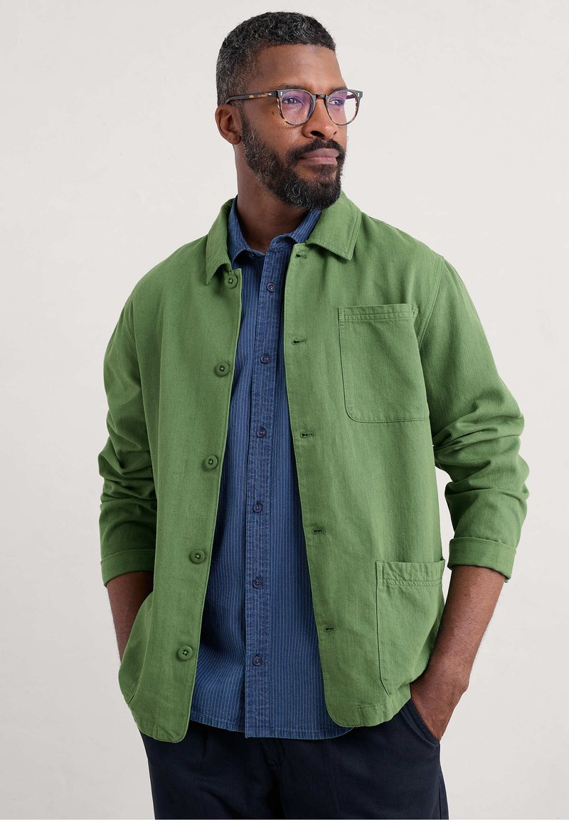 Green button-up shirt jacket in textured fabric, featuring a chest pocket and rolled sleeves, layered over a blue striped shirt.