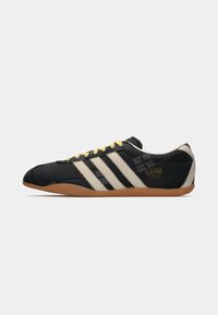 Black sneaker with three white stripes, yellow laces, and a textured upper. Features a gum sole and embroidered branding on the side.