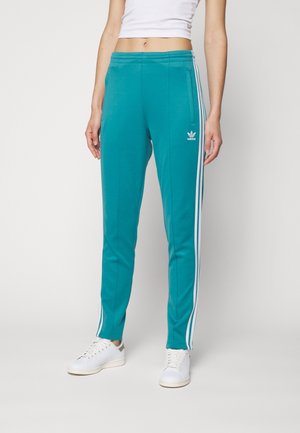 Frau trägt türkise Adidas-Trainingshose mit weißen Streifen und Logo, weiße Sneakers und ein weißes, bauchfreies Tanktop, steht vor einem einfarbigen Hintergrund.