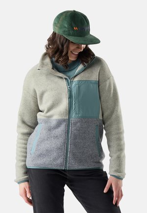Mujer con gorra verde y chaqueta polar de bloques de color con paneles en gris claro, azul y gris, sonriendo y mirando hacia abajo.