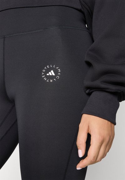 Leggings noirs en tissu extensible, avec un logo blanc en relief sur la cuisse et des coutures contrastantes mettant en valeur le design.
