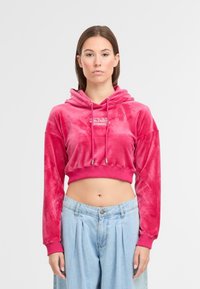 Gekürzte rosa Velourskapuzenjacke mit Kordelzug, die ein kontrastierendes Logo aufweist. Kombiniert mit hochgeschnittenen, hellen gefalteten Jeanshosen.