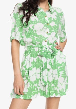 Kobieta ubrana w krótki kombinezon na guziki w zielono-białe kwiaty z wiązanym paskiem w talii, stoi z jedną ręką na biodrze.