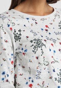 Top gris de manga larga con un patrón floral que presenta flores rojas, azules y verdes sobre una tela texturizada. Cuello redondo acanalado.