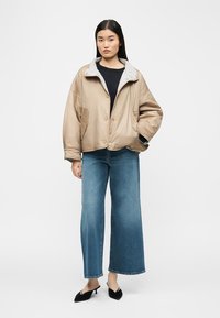 Γυναίκα που φοράει μπεζ φαρδιά ζακέτα με ριγέ φόδρα, μαύρο μπλουζάκι, μπλε τζιν παντελόνι με φαρδιά πόδια και μαύρες μυτερές γόβες, στέκεται μπροστά σε λευκό φόντο.