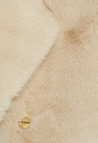 Tessuto di pelliccia sintetica beige con una consistenza morbida, caratterizzato da un bottone con il logo dorato "GUESS" a destra e una finitura liscia a sinistra.