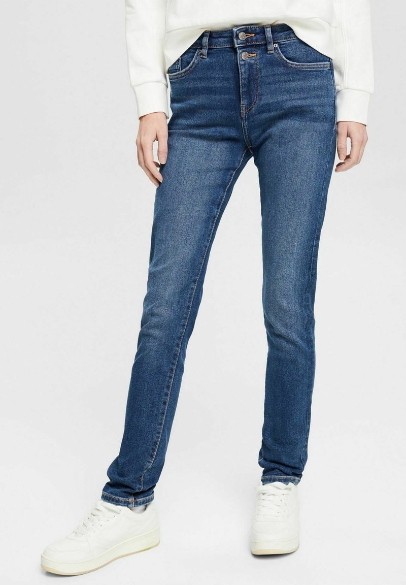 Jean slim bleu avec fermeture à double bouton, porté avec des baskets blanches et un haut blanc à manches longues, vu de la taille aux pieds.
