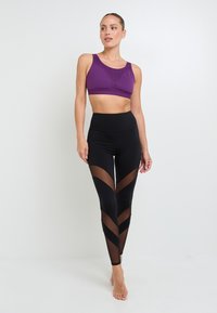 Donna in piedi a piedi nudi, vestita con un reggiseno sportivo viola e leggings neri a vita alta con pannelli in rete trasparente a forma di V.