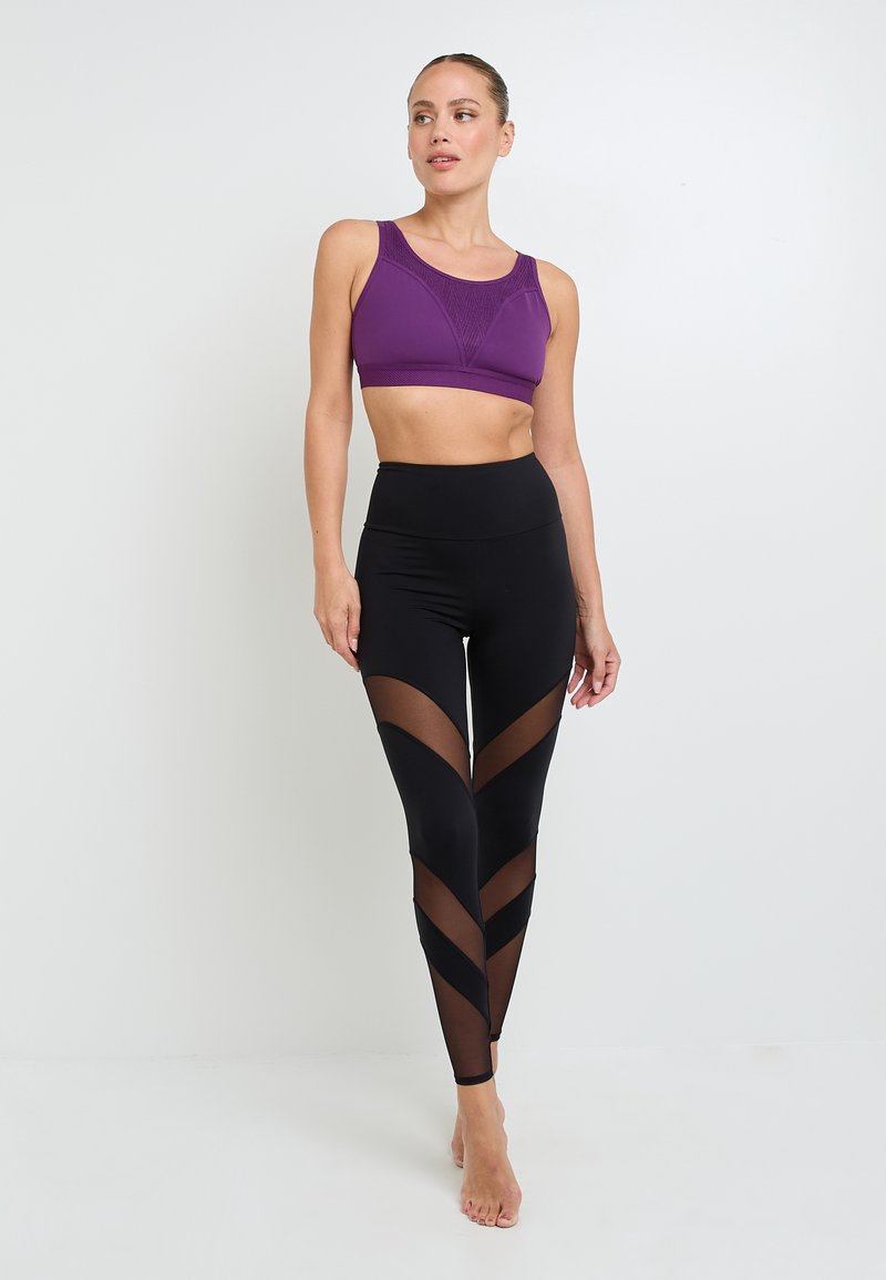 Donna in piedi a piedi nudi, vestita con un reggiseno sportivo viola e leggings neri a vita alta con pannelli in rete trasparente a forma di V.