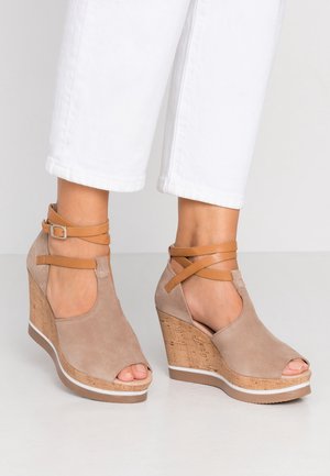 Beige ruskind kilehælssandaler med korkplatform, åbne tæer og mørkebrune læderstropper om anklen, båret med hvide bukser med opkrogede ben.