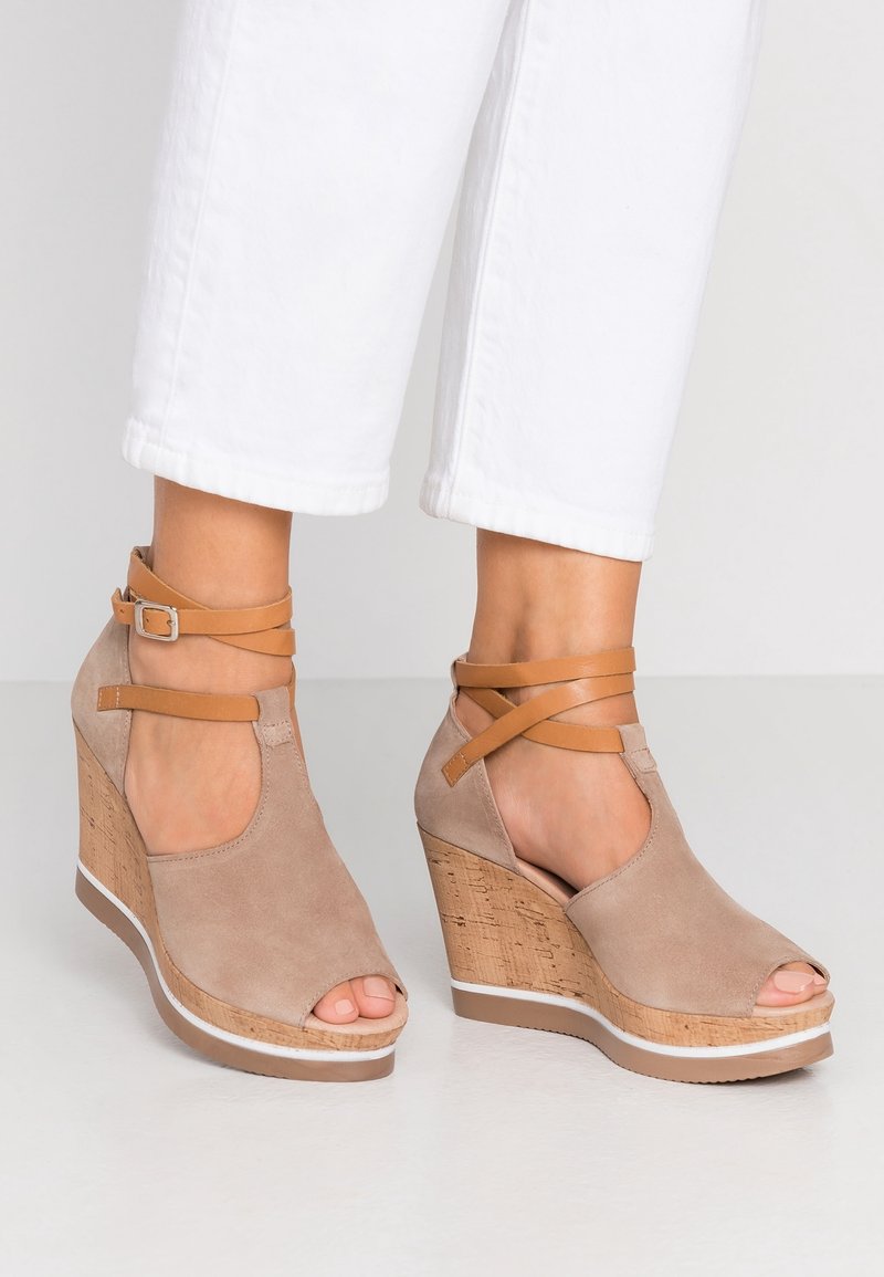 Sandales compensées en daim beige avec plateforme en liège, orteils ouverts, et brides de cheville en cuir marron, portées avec un pantalon blanc raccourci.