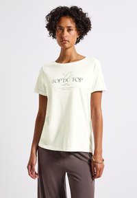Lys beige kortærmet t-shirt lavet af blødt stof, med mørkebrune bogstaver på forsiden. Løs pasform med rund hals og minimalistisk design.