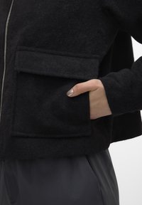 Musta villasekoitus takki, jossa on vetoketjusulku ja etuflanin tasku. Ominaisuuksina pehmeä, teksturoitu pinta ja istuvat hihat.