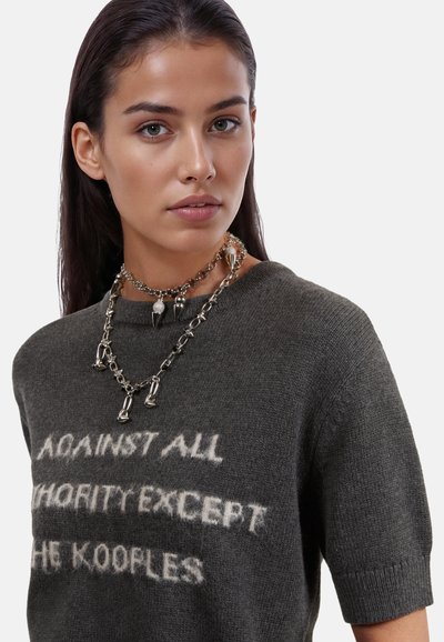 Grijze gebreide trui met korte mouwen toont tekst in het wit: "TEGEN ALLE AUTORITEIT UITGEZONDERD DE KOOPLES." Sieraden bestaan uit gelaagde zilveren kettingen en spikes.