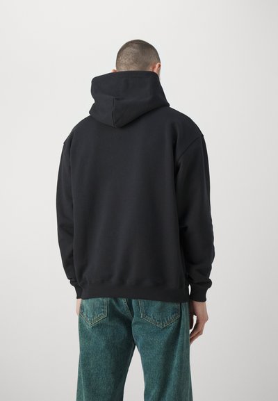 Schwarzer Kapuzenpullover mit lockerer Passform, gerippten Bündchen und Saum. Das Material wirkt weich und hat eine glatte Textur. Kombiniert mit grünen Jeans.