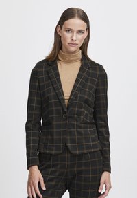 Blazer noir avec un motif à carreaux et des accents dorés, coupe ajustée, fermeture à un bouton et deux poches avant. Porté par-dessus un pull à col roulé beige.