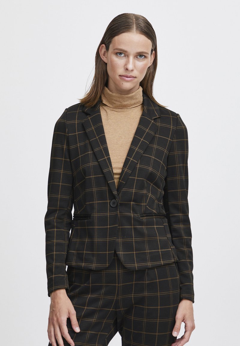 Blazer noir avec un motif à carreaux et des accents dorés, coupe ajustée, fermeture à un bouton et deux poches avant. Porté par-dessus un pull à col roulé beige.