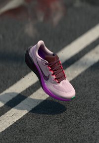 Zapatilla de running rosa claro con parte superior de malla, detalles en marrón oscuro y cordones rosa brillante. Cuenta con una mediasuela morada y una suela externa verde.