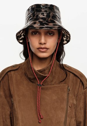 Vrouw draagt een bruine suèdeleren jas en een zwartbruine panterprint emmerhoed met een rode kinband tegen een witte achtergrond.
