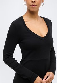 Γυναίκα που φοράει μαύρο μπλουζάκι με μακριά μανίκια και V-λαιμόκοψη, με κρίκους στα αυτιά, εμφανιζόμενη από τον λαιμό μέχρι τη μέση, μπροστά σε απλό φωτεινό φόντο.