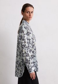 Femme portant un chemisier à manches longues bleu clair avec des motifs floraux sombres, se tenant de côté contre un mur blanc uni.