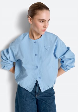 Haja hátrafésült nő világoskék, gombos kabátot visel elasztikus mandzsettákkal és sötétkék farmert, oldalra néz.