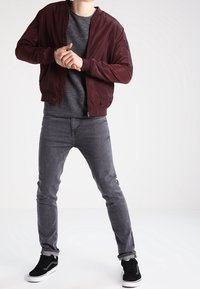 Blouson aviateur bordeaux, pull en maille gris, jean gris et baskets en daim noir. Le blouson est doté d'une fermeture éclair à l'avant et de poignets côtelés.