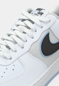 Weiße Nike Air Force 1 Schuhe mit grauem Mesh-Obermaterial, schwarzem Swoosh-Akzent und hellblauen Details. Merkmale sind flache weiße Schnürsenkel und eine perforierte Zehenbox.