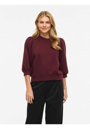 Jeune femme aux longs cheveux blonds portant un haut bordeaux à manches bouffantes et un pantalon large noir, debout devant un fond blanc.