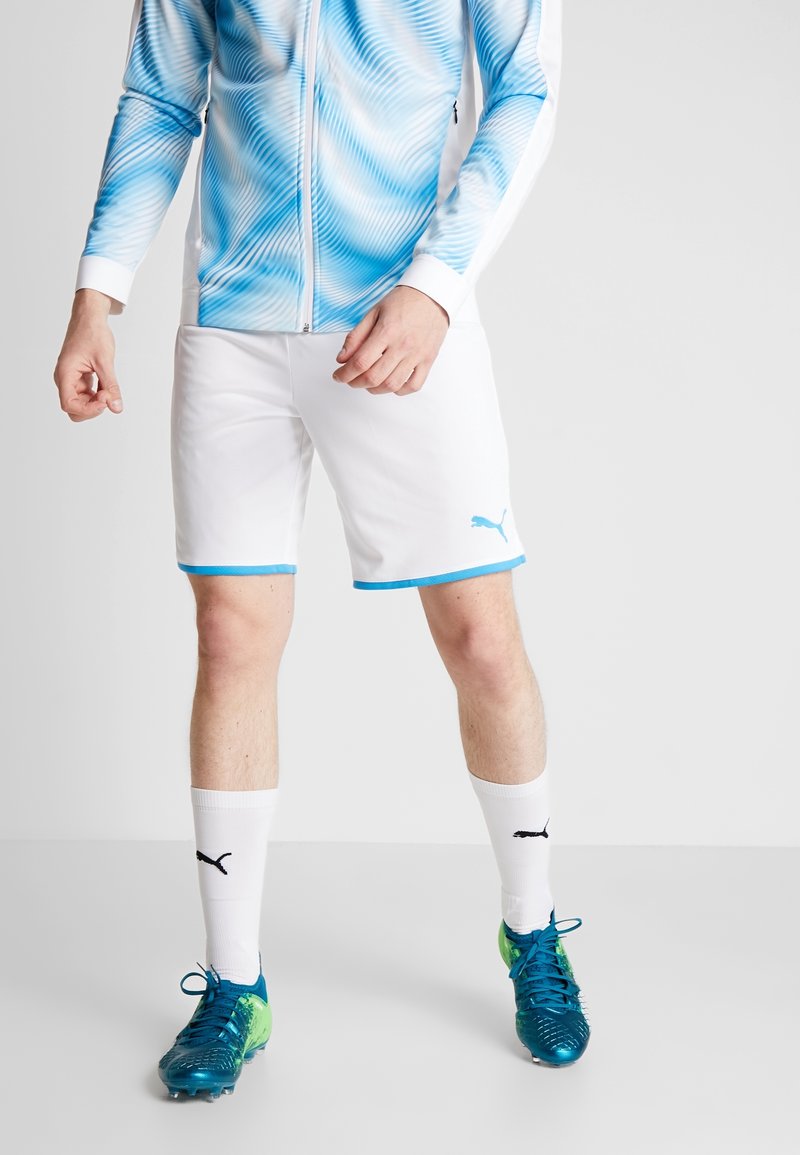 Shorts de sport blancs avec des bords bleus, associés à une veste zippée bleu et blanche à motifs, des chaussettes blanches et des chaussures de football bleu-vert.